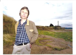 David Greygoose standing on a grassy, rocky shore with a lighthouse in the distance.